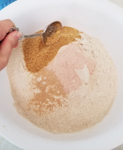A hand tips a tablespoon full of ingredients into a while bowl, on top of flour and other ingredients.