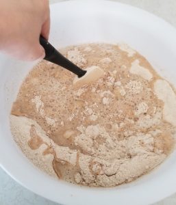 A hand mixes flour and dry ingredients together in a white bowl using a spatula.