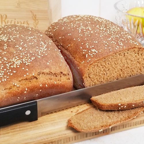 Two Brown Loafs of Bread sit on a cutting board. One loaf is sliced with the bread knife resting inbetween slices.