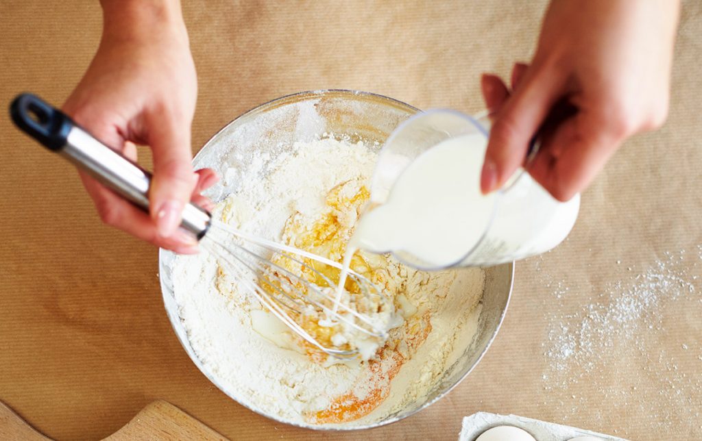 Women pouring out measured milk in a measuring cup into a mixing bowl with flour and egg.